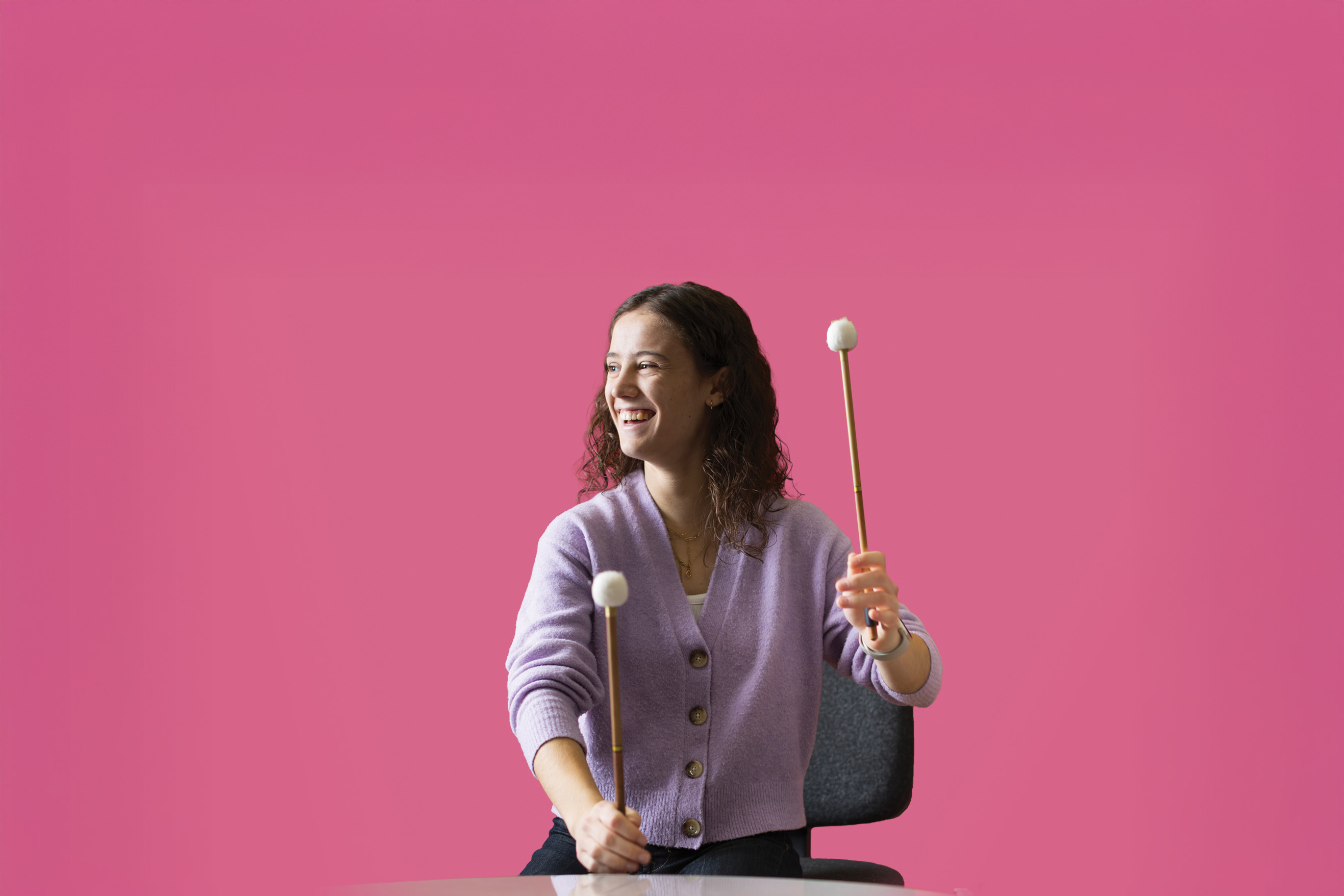 A woman playing the timpani and looking to the side, with a deep, rose pink background.